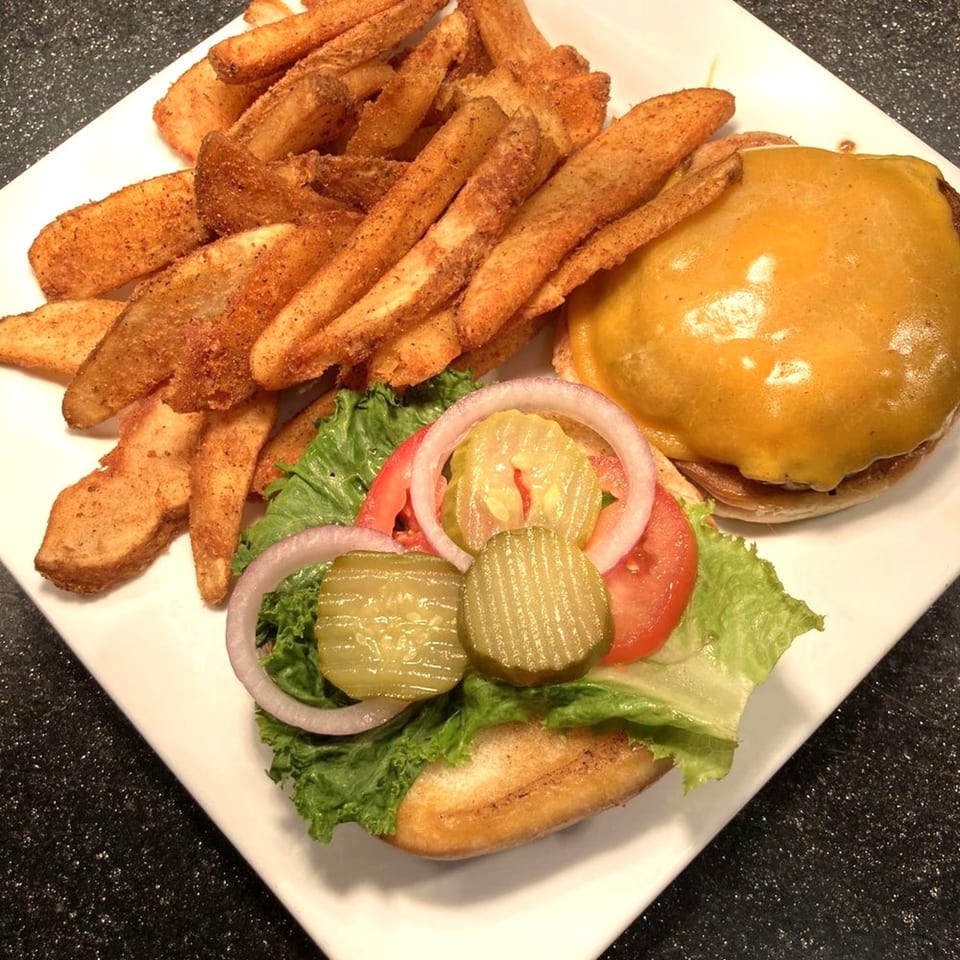 Cheeseburger with lettuce, tomato, onion, pickles, and a side of seasoned potato wedges on a white plate.