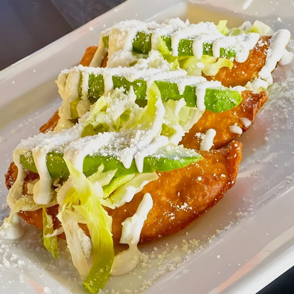 Three golden-fried Mexican empanadas topped with lettuce, avocado slices, crema, and cotija cheese.