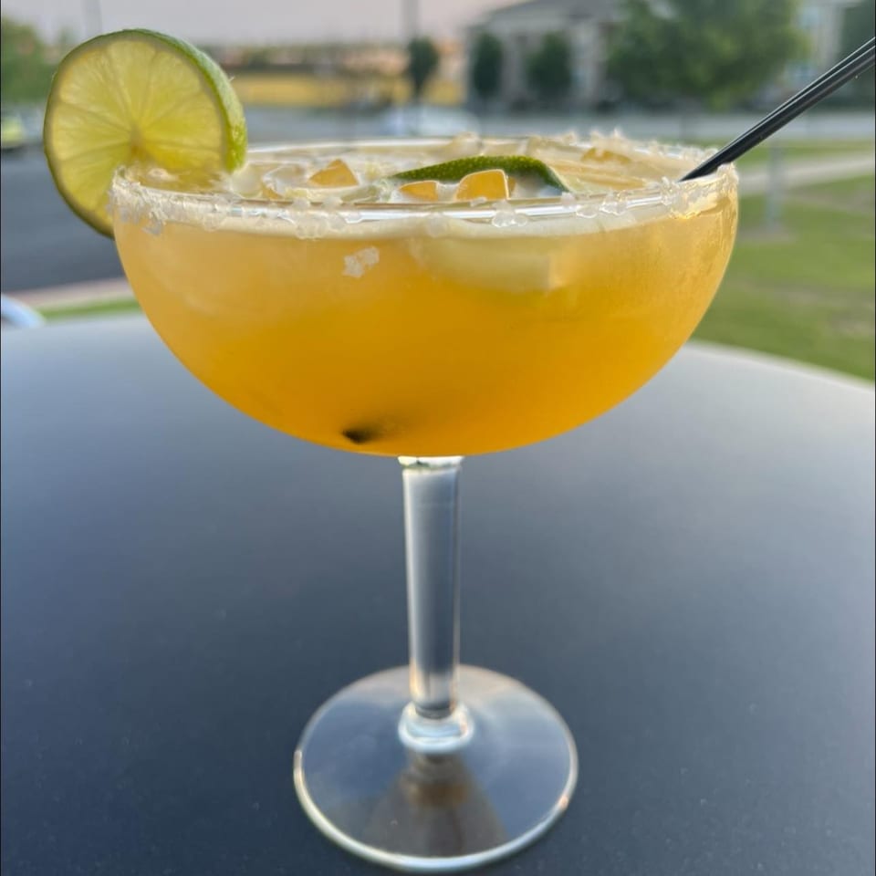 A vibrant yellow margarita with a salted rim, lime slice, and black straw, outdoors at Angus Grill & Cantina.