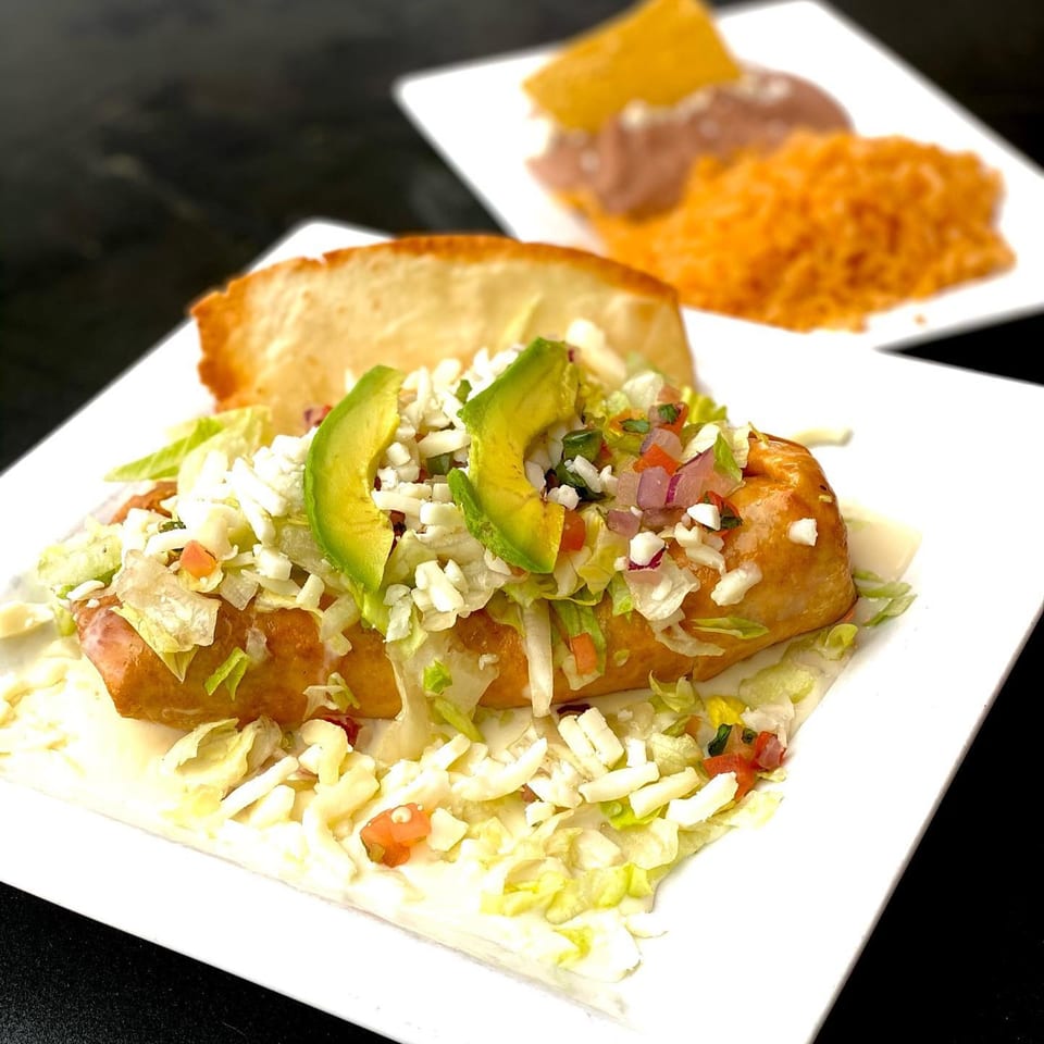 A golden-brown chimichanga topped with lettuce, cheese, pico de gallo, and avocado slices, with rice and beans in the background.