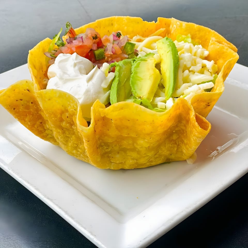 A vibrant Mexican taco salad in a crispy tortilla bowl, topped with pico de gallo, sour cream, avocado, and cheese.